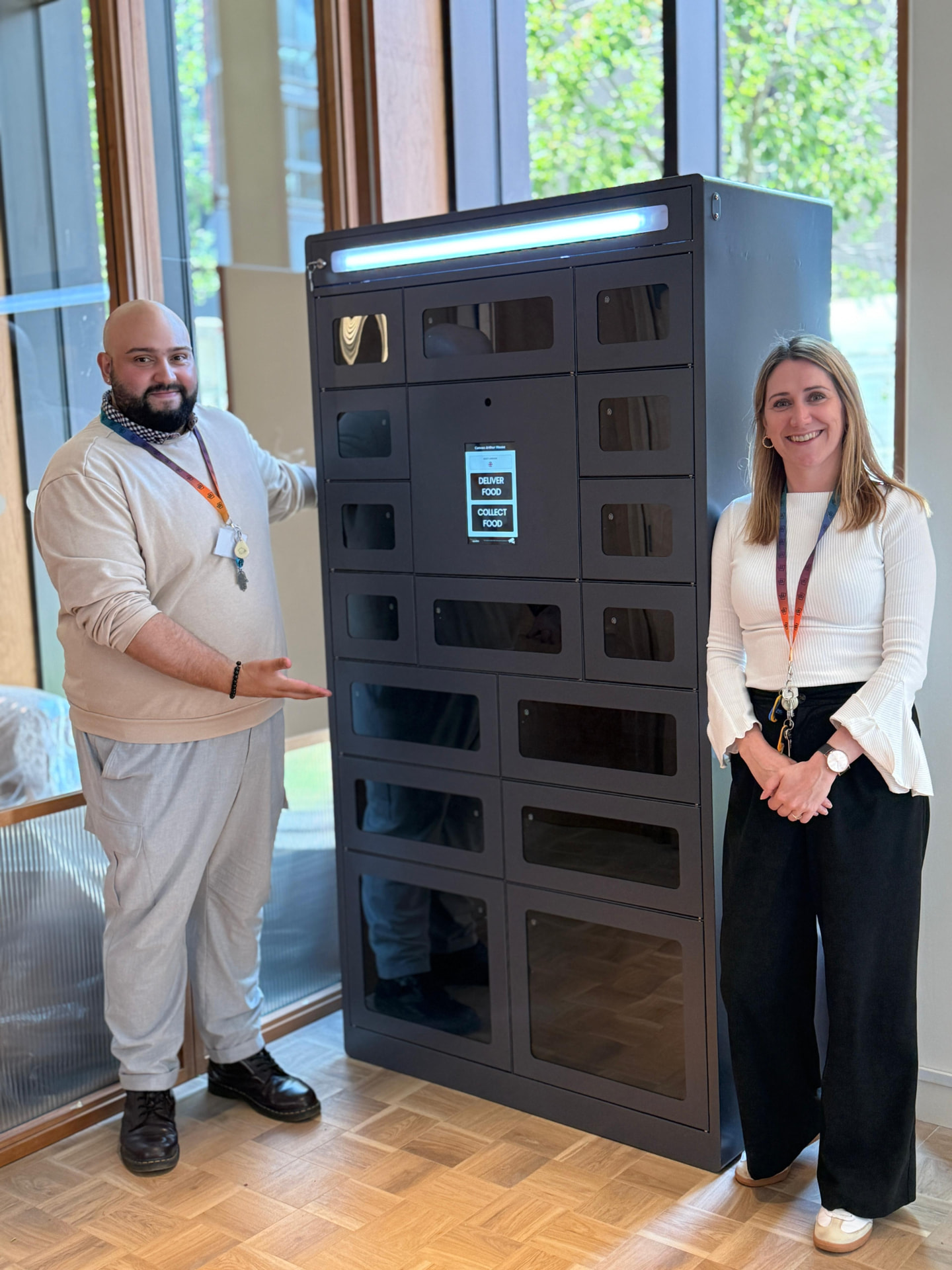 Two locker business owners standing beside a food delivery and collection locker in a bright building lobby